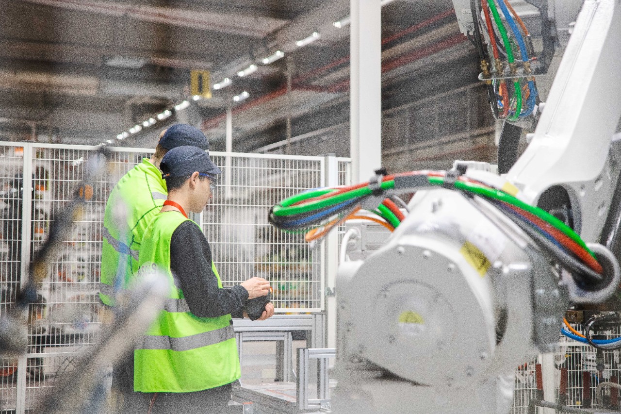 Praktikanter tillsammans med medarbetare står i en robotcell och kollar på en handdator