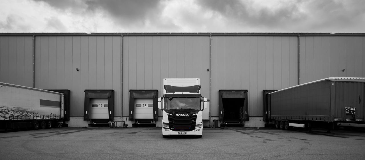 An electric Scania 25 P, positioned centrally in front of a warehouse loading dock, with loading bays and trailers on either side.