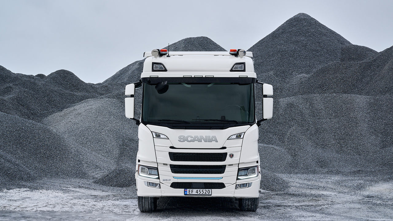 A white electric Scania 25 P parked in a gravel pit, with piles of gravel behind it.