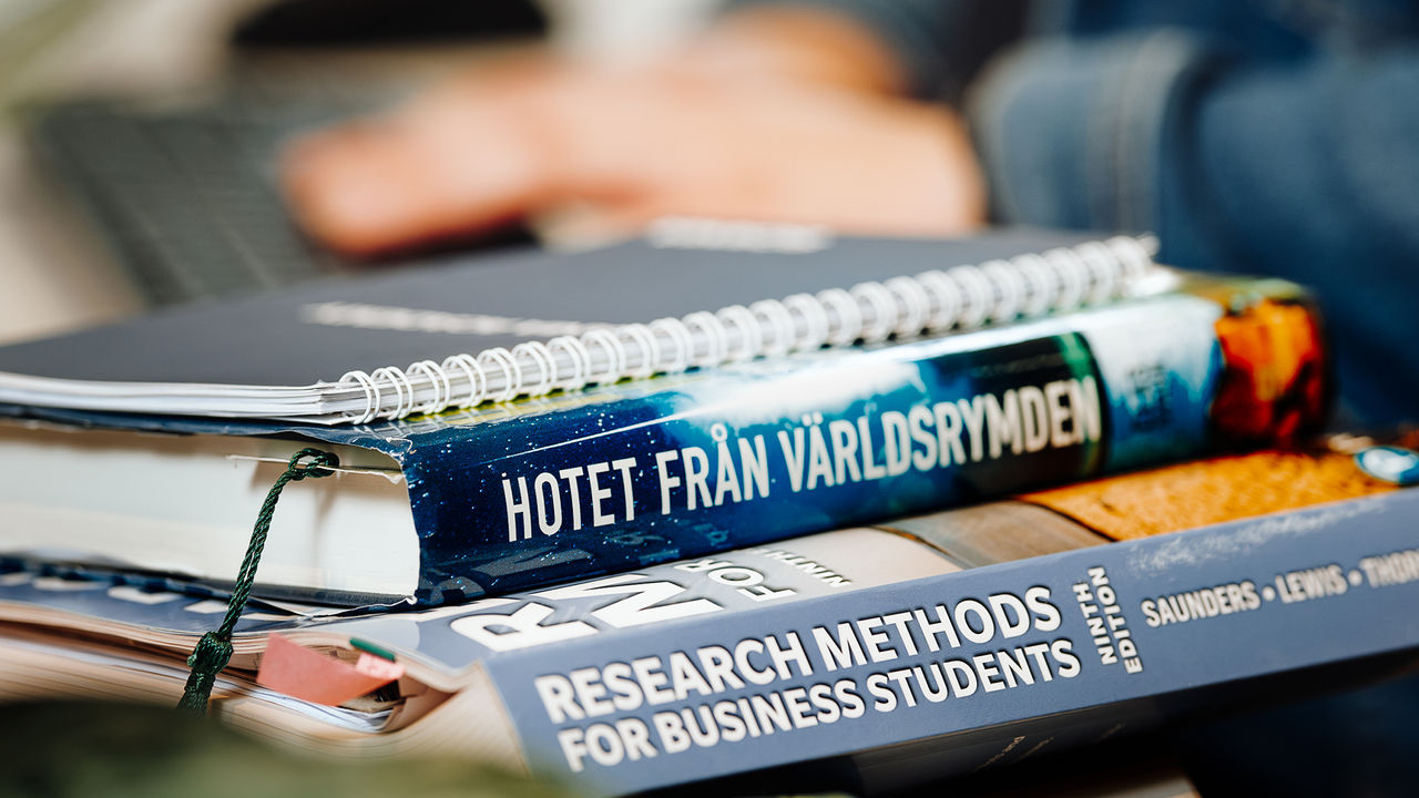 A stack of books and notebooks on a desk. One book is titled “Hotet från världsrymden” and another “Research Methods for Business Students.” In the background, Evy Svensson, Enterprise Architect at Scania is typing on a laptop.